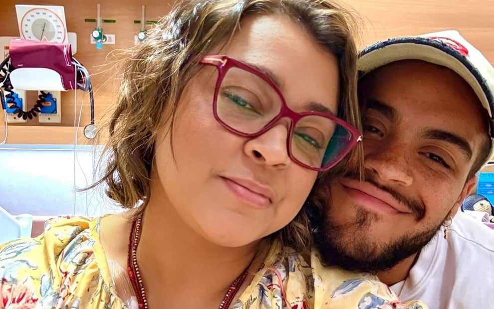 Preta Gil e o filho, Francisco Gil; eles dão um leve sorriso no hospital. A artista usa um óculos de aros vermelhos e uma blusa amarela; já o cantor está com uma blusa branca e um boné da mesma cor