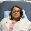 Mulher deitada em poltrona hospitalar, usando óculos e agasalho cinza, com acesso médico no peito ligado a equipamento de infusão.