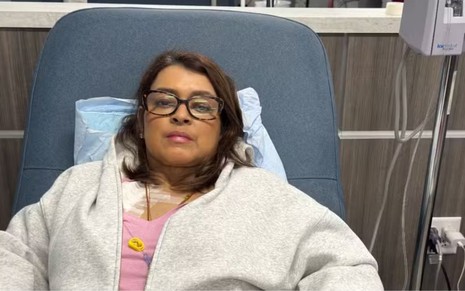 Mulher deitada em poltrona hospitalar, usando óculos e agasalho cinza, com acesso médico no peito ligado a equipamento de infusão.