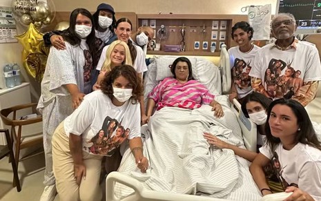 Preta Gil, deitada em um leito hospitalar, está cercada de pessoas; elas usam máscaras no rosto