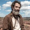 Rafael Sieg está com a barba longa, uma uma camiseta branca (mas com aspecto sujo) e um casaco marrom por cima; ao fundo há uma paisagem do sertão nordestino com céu azul e nuvens brancas