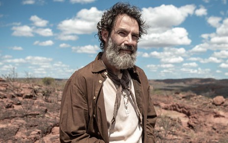 Rafael Sieg está com a barba longa, uma uma camiseta branca (mas com aspecto sujo) e um casaco marrom por cima; ao fundo há uma paisagem do sertão nordestino com céu azul e nuvens brancas