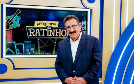 Ratinho sorridente em frente à logo do Programa do Ratinho, usando um terno azul e uma camisa branca, além de óculos de grau com hastes azuis