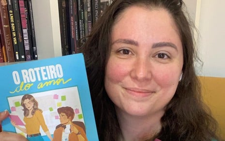 Ray Tavares sorri e segura o livro  O Roteiro do Amor nas mãos