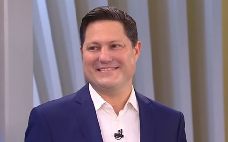 Homem de terno azul e camisa branca sorri em estúdio com fundo de painéis verticais em tons claros. Usa microfone de lapela.