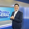 Apresentador do Balanço Geral SP sorri no estúdio, de terno azul-marinho e mãos entrelaçadas.