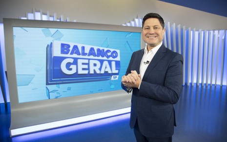 Apresentador do Balanço Geral SP sorri no estúdio, de terno azul-marinho e mãos entrelaçadas.