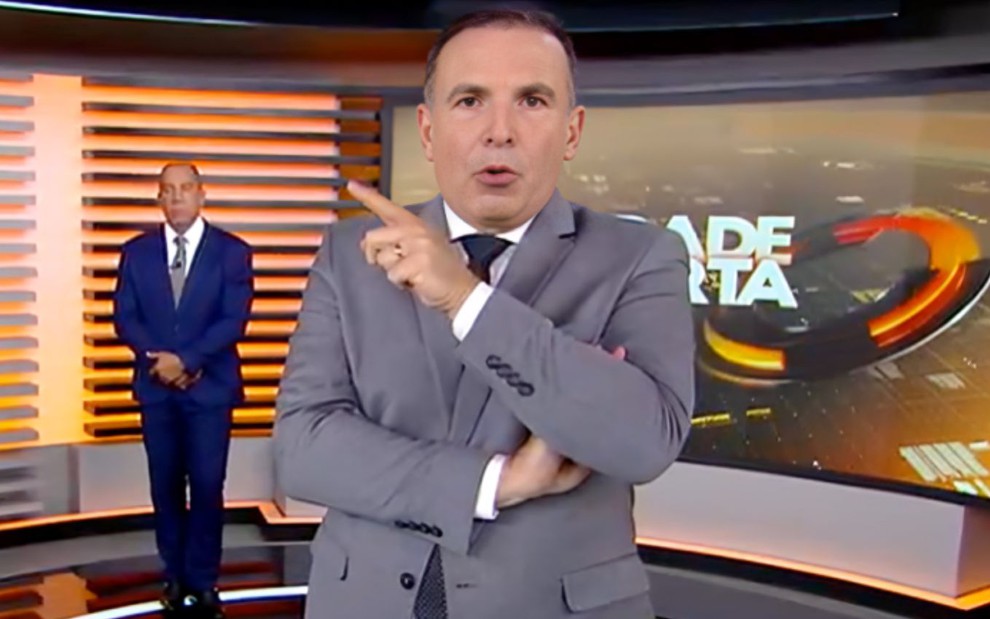 Dois apresentadores em estúdio de telejornal; um ao fundo sério, outro à frente gesticulando com expressão firme.