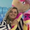 Mulher loira, sorrindo, veste blusa listrada preto e branco e segura uma cobra de pelúcia rosa e amarela com chapéu rosa. Ao fundo, um painel azul.