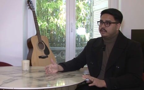 Com bigodinho e óculos de grau, um homem de casaco e blusa de gola rolê está em uma sala com um violão ao fundo