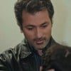 Feyyaz Duman caracterizado como Arif, olha para baixo com sorrido discreto em cena de Força de Mulher