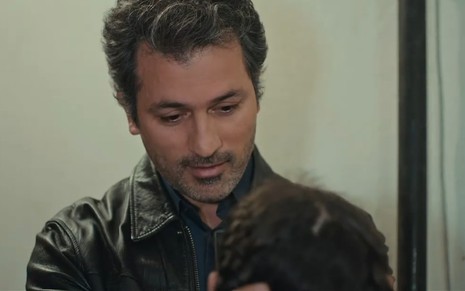 Feyyaz Duman caracterizado como Arif, olha para baixo com sorrido discreto em cena de Força de Mulher