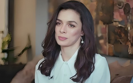 Mulher de cabelos longos e escuros, veste camisa branca e usa brinco de pérola. Ela está em ambiente interno, com expressão serena.