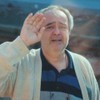 Şerif Erol tem expressão triste e se despede de uma mulher, que o observa por uma janela de vidro