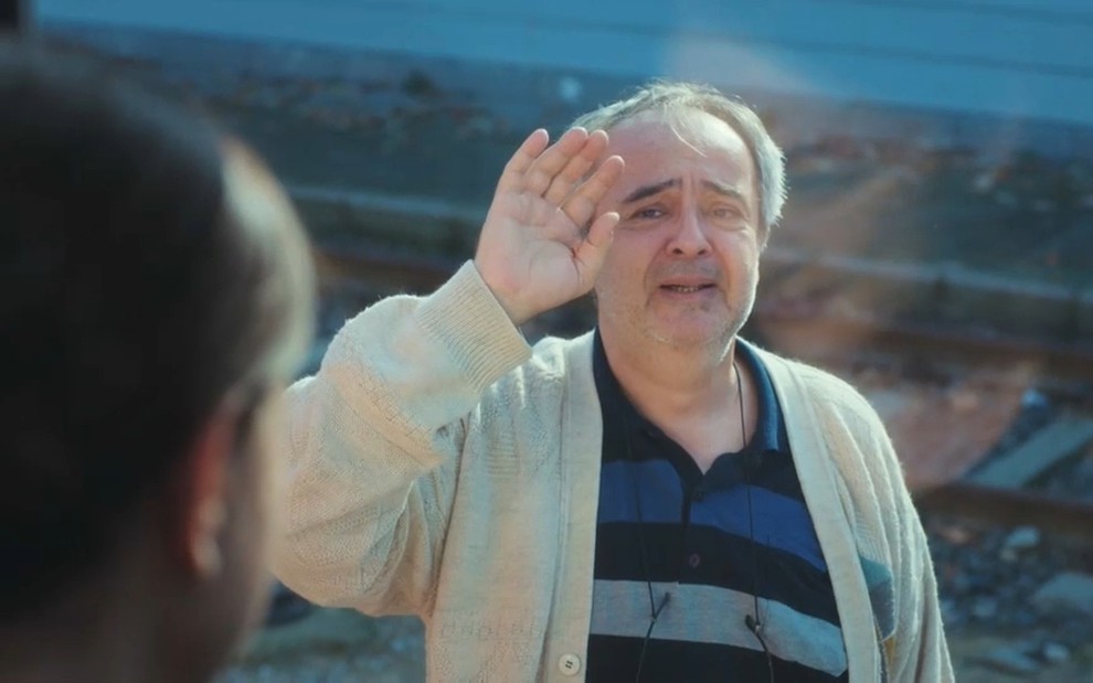 Şerif Erol tem expressão triste e se despede de uma mulher, que o observa por uma janela de vidro