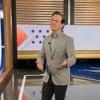 Homem em estúdio de TV, vestindo blazer cinza e camisa social, faz pose descontraída com expressão confiante.