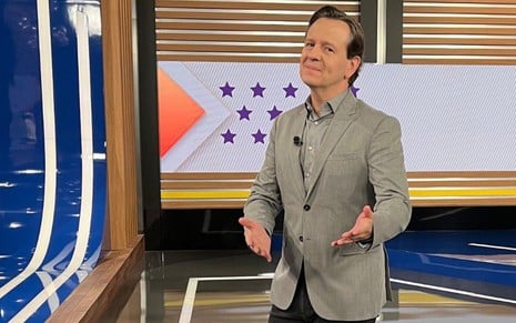 Homem em estúdio de TV, vestindo blazer cinza e camisa social, faz pose descontraída com expressão confiante.
