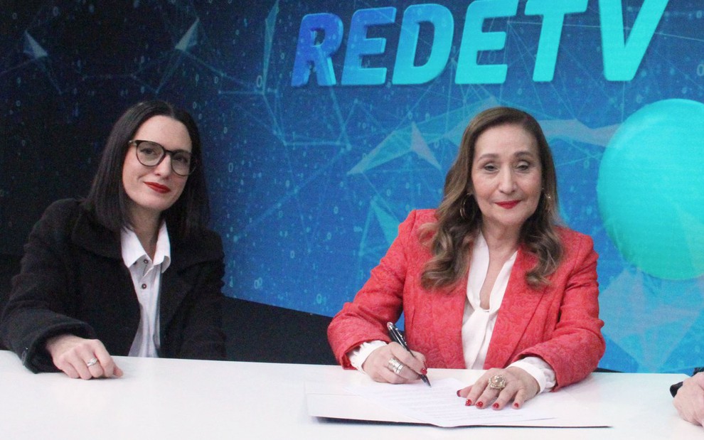 Andrea Dallevo e Sonia Abrão posam para foto enquanto a segunda segura uma caneta em cima de um documento