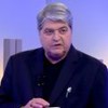Homem de cabelo grisalho veste terno escuro em estúdio com luzes azuladas e barras verticais decorativas ao fundo.