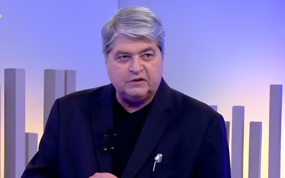Homem de cabelo grisalho veste terno escuro em estúdio com luzes azuladas e barras verticais decorativas ao fundo.
