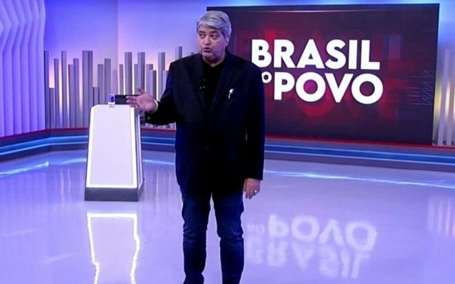 Homem de cabelos grisalhos, vestindo blazer preto e calça jeans, fala em estúdio moderno com o letreiro "BRASIL DO POVO" ao fundo.
