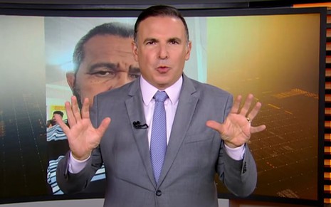 Apresentador de terno cinza fala com as mãos erguidas diante de telão com imagem de homem barbudo ao fundo.