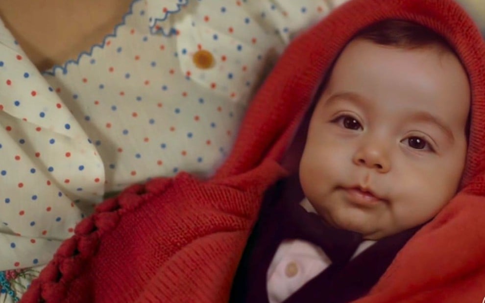 Bebê de olhos escuros é mostrado de perto, enrolado em manta vermelha no colo de uma pessoa com vestido estampado.