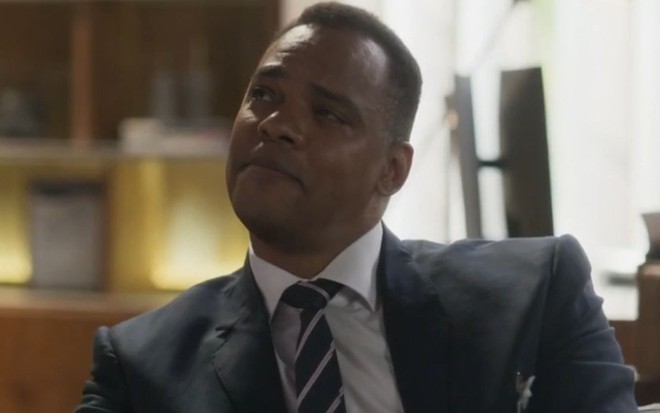Rocco Pitanga usa terno e sorri discretamente em cena da novela Cara e Coragem (2022)