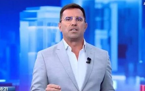 O jornalista Rodrigo Bocardi, de paletó cinza-claro, camisa branca sem gravata e óculos de armação transparente, apresenta ao vivo no estúdio com fundo azul.