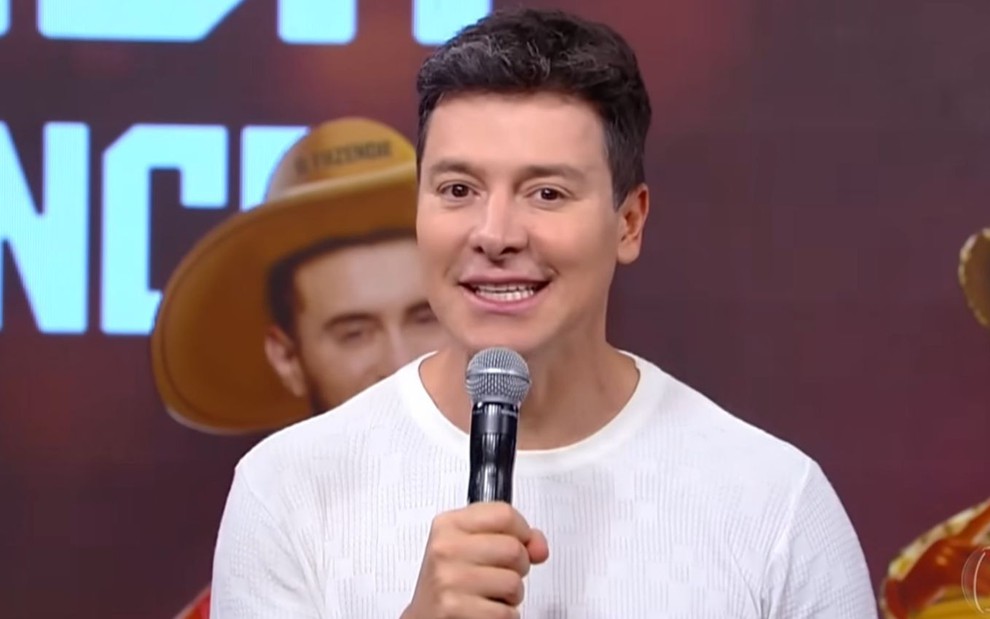 Homem de cabelo curto e camisa branca segura um microfone, com um fundo de palco mostrando um chapéu de cowboy e texto ao fundo.