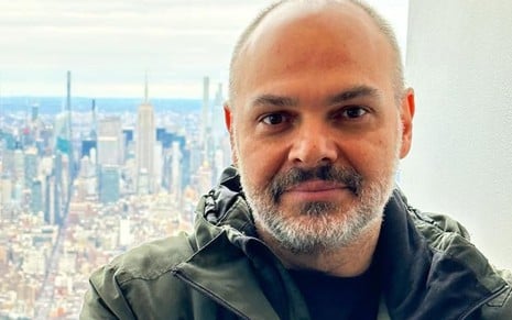 Homem calvo com barba curta e jaqueta verde, em frente a um panorama urbano com arranha-céus, incluindo o Empire State Building.