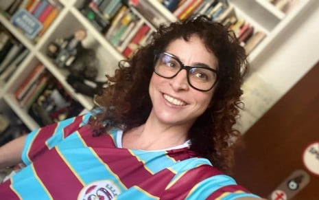 Rosane Svartman usa uma camiseta listrada em azul e rosa; ela sorri para a câmera