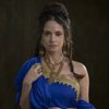 Rosanne Mulholland está com cabelo semi preso, usa um vestido azul de uma manga só e vários colares dourados