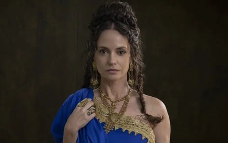 Rosanne Mulholland está com cabelo semi preso, usa um vestido azul de uma manga só e vários colares dourados