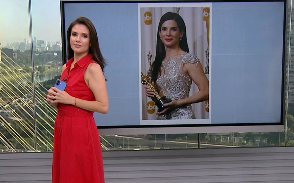 Sabina Simonato imitando Sandra Bullock segurando um Oscar; a imagem da atriz de Hollywood aparece no telão ao fundo