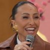 Sabrina Sato sentada no sofá Encontro, com um casaco de couro marrom, cabelo preso, segurando o microfone e rindo