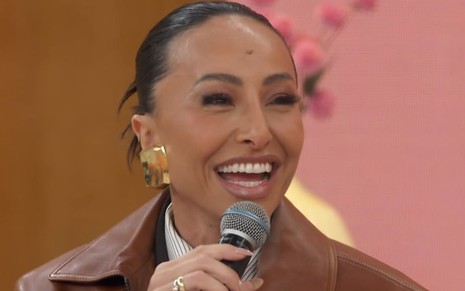 Sabrina Sato sentada no sofá Encontro, com um casaco de couro marrom, cabelo preso, segurando o microfone e rindo