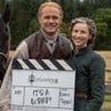 Sam Heughan e Caitriona Balfe seguram uma claquete no set de Outlander; um cavalo preto aparece atrás deles