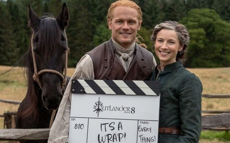 Sam Heughan e Caitriona Balfe seguram uma claquete no set de Outlander; um cavalo preto aparece atrás deles