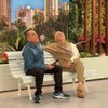 Carlos Alberto de Nóbrega e humorista sentam em banco de praça, rindo juntos durante gravação em estúdio.