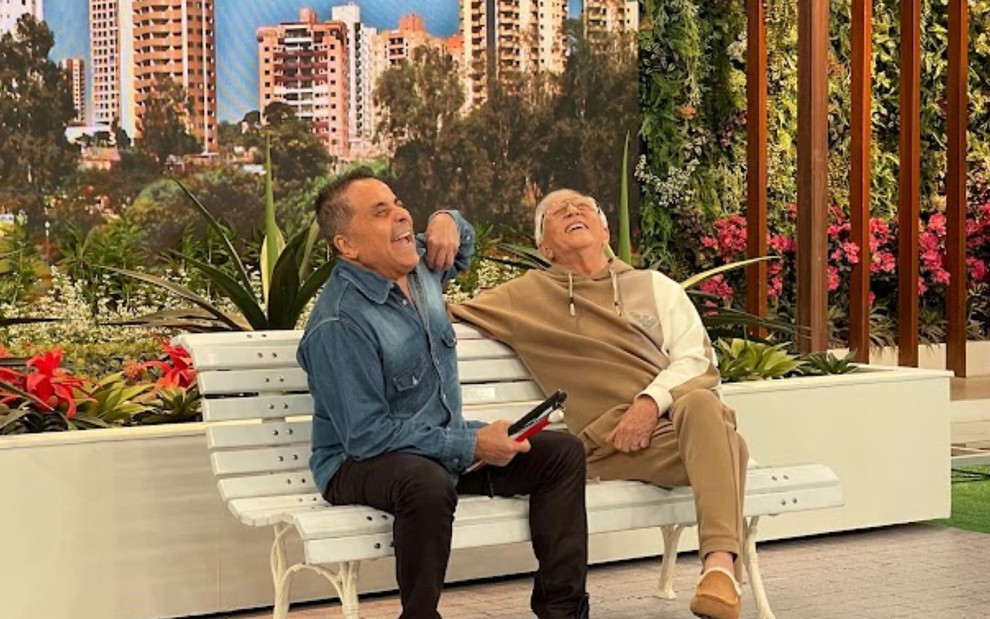 Carlos Alberto de Nóbrega e humorista sentam em banco de praça, rindo juntos durante gravação em estúdio.