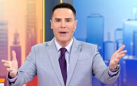 Luiz Bacci está de braços abertos, em tom de súplica, no cenário do Alô, Você