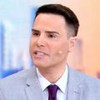 Homem de terno cinza claro e gravata roxa fala com microfone auricular em estúdio com fundo desfocado em tons azul e laranja.