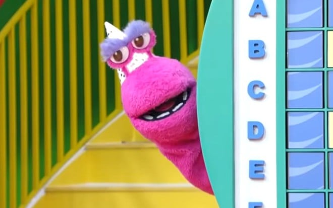 Fantoche rosa com sobrancelhas roxas e chifre branco aparece sorrindo atrás de um painel colorido com letras do alfabeto.