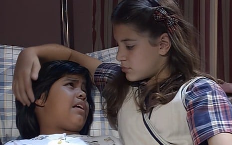 Giselle Medeiros está deitada na cama e chora, enquanto Renata Del Bianco faz carinho em sua cabeça em cena de Chiquititas