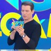 Homem de camisa preta apresenta programa de TV, segurando microfone e cartão colorido com fundo azul vibrante e letras grandes.