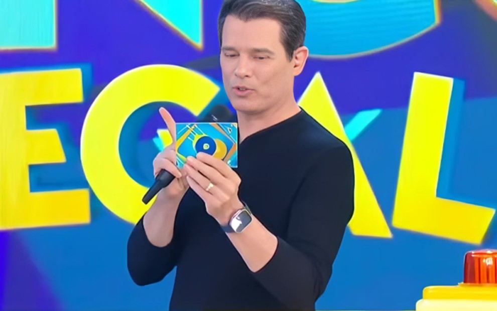 Homem de camisa preta apresenta programa de TV, segurando microfone e cartão colorido com fundo azul vibrante e letras grandes.