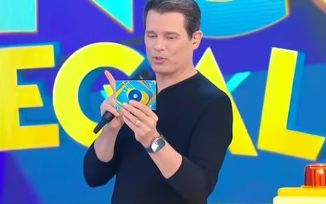 Homem de camisa preta apresenta programa de TV, segurando microfone e cartão colorido com fundo azul vibrante e letras grandes.