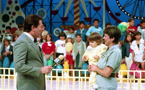 Silvio Santos conversa com uma mãe que segura uma criança no colo no cenário do Domingo no Parque