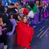 Mulher com microfone preso à roupa vermelha se destaca na plateia de programa; outras pessoas riem e se abaixam nas cadeiras lilás.